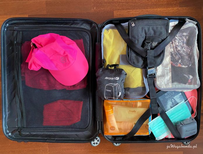 ubrania w workach, składany plecak, poduszka podróżna dmuchana i czapka spakowane do małej walizki