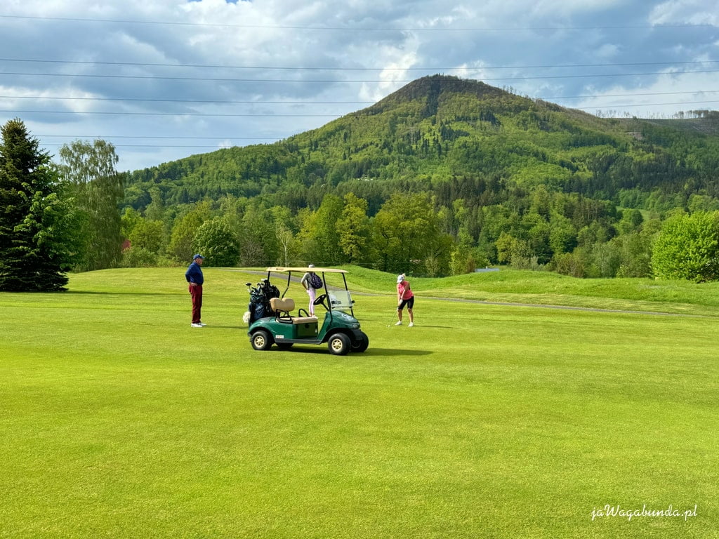 wózek golfowy, kobieta gra w golfa, mężczyzna stoi obok, w tle góry