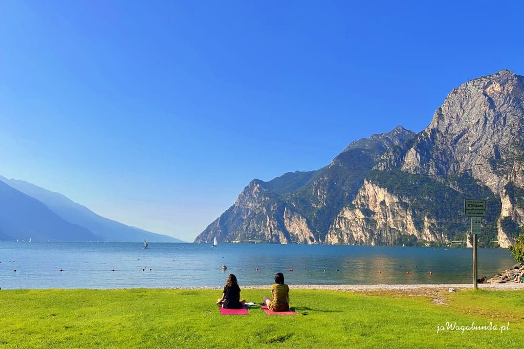 dwoje ludzi siedzi na brzegiem jeziora do którego spadają wysokie góry